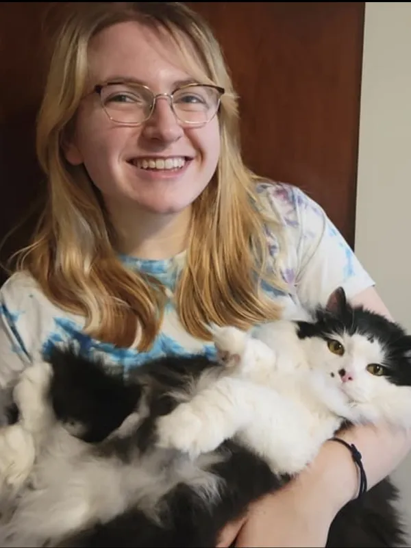 Maddy McCain (they are a blonde haired person, with glasses, holding a black and white cat while smiling)